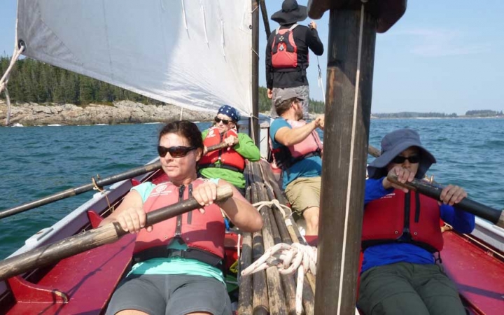 Four people sit and row a sailboat on calm blue water. In the background, there is a rocky and tree-lined shore.